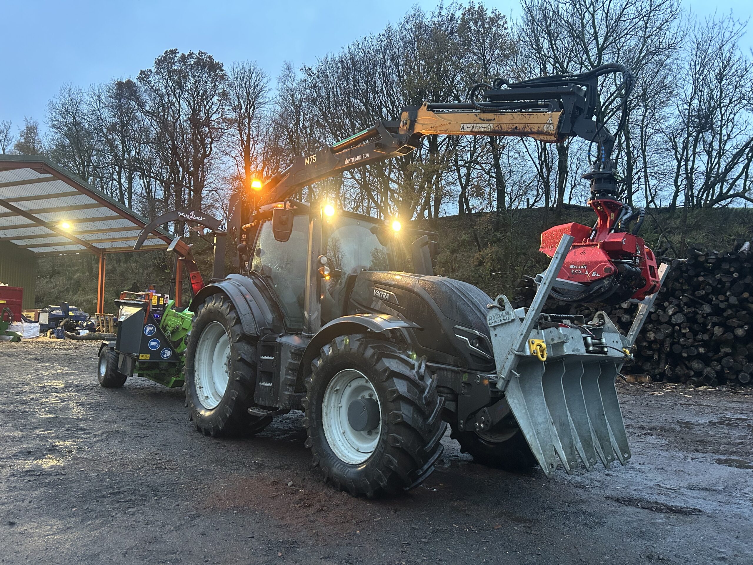 A tractor fitted with a timber grapple crane and wood chipper working in woodland.