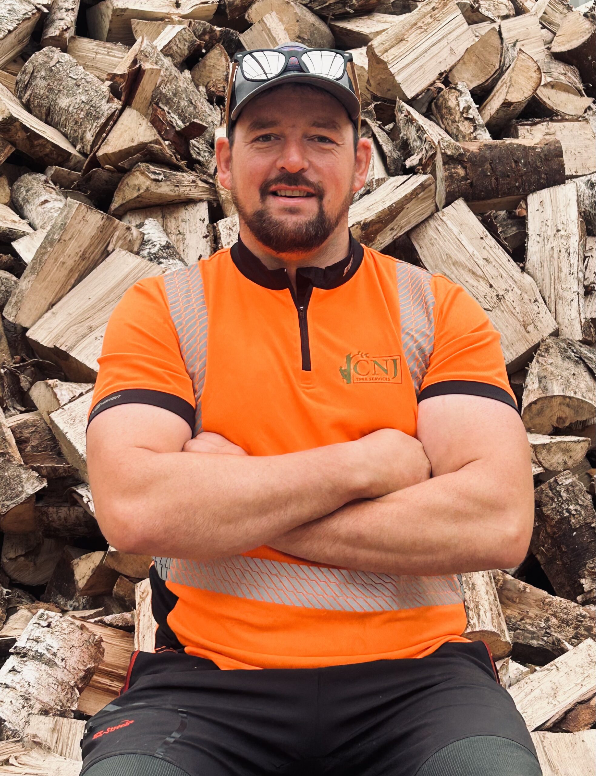 A tree surgeon standing in front of a large firewood log pile.